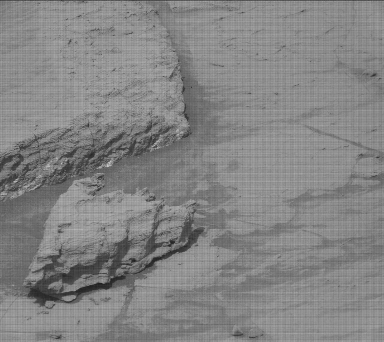 A grayscale photo from the Martian surface shows a barren, rocky landscape featuring a jagged, heavily striated rock formation resting on a flat, cracked surface beside a larger rocky ledge.